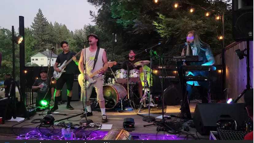 Outdoor band performance with guitarist, drummer, and keyboardist under string lights.